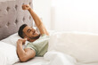 © Prostock-studio - A young man sits up in bed, stretching his arms and smiling in a bright, sunny bedroom. The soft white sheets and sunlight create a warm, cheerful atmosphere.