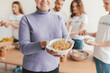 © Prostock-studio - A woman smiles while holding a bowl of colorful food in a kitchen where friends are engaged in cooking. The atmosphere is friendly and lively as they share food.
