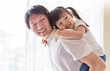 © paulaphoto - Portrait of smile father giving his young daughter a piggyback ride indoors, Warm and joyful family bonding moment at home. Father’s day
