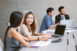 © edward_indy - Two Asian colleagues discuss work at a laptop while smiling, with charts and documents on the table. In the background, two teammates focus on their tasks and productive office environment.