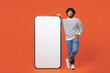 © ViDi Studio - Full body young smiling happy Indian man wear striped sweatshirt casual clothes stand near big huge blank screen mobile cell phone smartphone with mockup area isolated on plain red orange background.