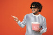 © ViDi Studio - Young happy Indian man he wears striped sweatshirt casual clothes 3d glasses watch movie film hold bucket of popcorn in cinema point aside isolated on plain red orange background. Lifestyle concept.