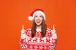 © ViDi Studio - Merry young woman wear red warm Christmas sweater Santa hat posing point index finger overhead indicate on area mock up isolated on plain orange background. Happy New Year celebration holiday concept.
