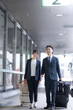 © kapinon - ビジネス出張でスーツケースを引く会社員の笑顔の移動シーン　全身
