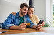 © baranq - Young joyful couple doing online shopping with mobile phone and credit card sitting at dinning table at home