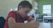 © vectorfusionart - Writing male student in red shirt leaning in at classroom desk, holding pencil, backpack visible