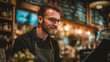 © Maryna - A relaxed barista with glasses and a beard smiles while reviewing a menu in a cozy cafe, surrounded by warm lighting and a cheerful atmosphere.