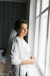 © Anhelina  - The bride in a white robe stands by the window holding a cup of coffee. She smiles softly, enjoying a calm moment before the wedding. A warm and peaceful morning atmosphere in a cozy room.