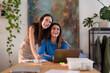 © Studio Marmellata - Two young women are smiling and working together at a desk with laptops and notebooks in a bright, modern room.
