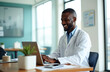 © miss irine - Smiling doctor in white lab coat uses laptop at desk in modern clinic. Black man works on computer reviewing patient data, happy male doctor smiles during online consultation with positive attitude.