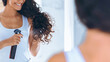© Prostock-studio - A woman with curly hair is applying hair product from a spray bottle while looking at herself in the mirror. The setting is a bright bathroom, highlighting her focus on hair care.