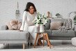© Pixel-Shot - Beautiful young happy African-American woman with vase of bouquet of white lily flowers sitting on sofa at home