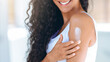 © Prostock-studio - A woman with curly hair is applying lotion to her shoulder, smiling enjoyably. The scene is bright and cheerful, suggesting a skincare routine during daytime.