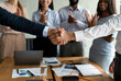 © Prostock-studio - Two men shake hands in a modern office after finalizing an agreement. In the background, diverse team members applaud in celebration of their successful collaboration.