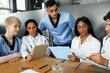 © Prostock-studio - Professional international team of doctors men and women in uniforms sitting at table, using digital tablet, taking notes at medical charts, having morning breefing, clinic interior, closeup
