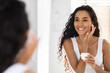 © Prostock-studio - A woman smiles at her reflection while applying cream to her face in a well-lit bathroom. She has long curly hair and wears a simple white tank top, showcasing her skincare routine.