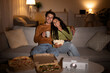 © Prostock-studio - A couple sits on their couch enjoying a movie night at home. They smile while holding coffee and popcorn, surrounded by pizza boxes and a projector, creating a cozy atmosphere together.