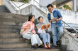 © bongkarn - Asian man student standing showing tablet to woman friends sitting on building stairs in university.
