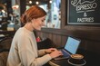 © Samina - Young woman with red hair focused on her laptop in a cozy cafe enjoying a coffee and pastries