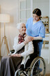 © AnnaStills - Senior Caucasian woman sitting in wheelchair with disability, smiling while young adult Hispanic female nurse standing behind her, gently placing hands on shoulders, both interacting indoors
