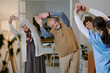 © AnnaStills - Group of senior Caucasian man and women following young adult female instructor stretching arms during fitness class indoors, all participants standing and engaging in physical exercise together