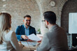 © Novak - Three business professionals sitting at a table in a modern office, engaged in a lively conversation. A man in a blue blazer is speaking enthusiastically
