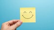 © Albaloshi - A hand holding a yellow happy smile face on blue background post-it note with a joyful expression, perfect for a motivational message or a quick moment of encouragement.
