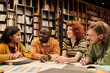 © Mediaphotos - Group of diverse young people sitting at table in library discussing project, smiling and making notes, multiethnic men and women collaborating on academic assignment together