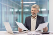 © Liubomir - Experienced professional man sitting at a desk with a laptop, smiling while confidently reviewing important business paperwork in a bright corporate environment
