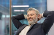 © Liubomir - Mature executive man smiling while relaxing and leaning back with hands behind his head, celebrating achievement and feeling successful in a modern office environment