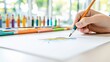 © Kairi - A hand is drawing with a colored pencil on a white sheet of paper, surrounded by other colored pencils. The scene is indoors with natural light.