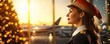 © VictoriaEmerson - Professional woman pilot wearing santa hat and uniform, looking out airport window, seeing airplanes, celebrating holiday travel season