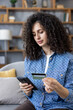 © Liubomir - Woman holding a credit card and smartphone at home, focused on entering payment information for convenient online shopping and e-commerce transactions