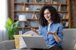 © Liubomir - Young woman on a cozy living room sofa gesturing and smiling while participating in a laptop video conference for remote work, online teaching or virtual meetings