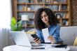 © Liubomir - Woman holding a receipt and smartphone, comparing expenses while managing her budget at home, experiencing financial stress and worry over incoming bills