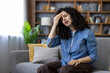 © Liubomir - Young woman suffering from severe headache or stress, sitting on a comfortable gray couch in a modern living room, eyes closed and holding her temples experiencing discomfort