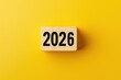 © oksa_studio - Wooden block displaying the year 2026 on a vibrant yellow background, providing ample copy space for text