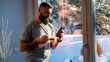© DusanJelicic - Bearded man wearing casual clothes enjoying a cup of coffee and using smartphone by the window in a snowy winter landscape, sunlight illuminating the scene