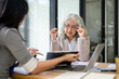 © bongkarn - Old asian businesswoman holding glasses talking to employee office worker at table in company room.