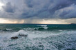 © Laura Pashkevich - Dramatic view of strong wind and high waves crashing over rocks under a dark, brooding storm sky. Intense marine landscape with a visible rain squall on the horizon.