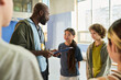 © pressmaster - Black man coach holding clipboard talking to diverse group of teenagers including Asian girl and Caucasian boy in sports facility during team meeting, other teens listening