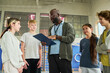 © pressmaster - Black man instructing diverse group of teenagers in gymnasium, holding clipboard and engaging students in discussion, teenagers listening attentively during sports activity