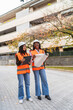© Koldo_Studio - Two smiling women engineers reviewing architectural plans on a job site, wearing hard hats and safety vests, symbolizing teamwork and diversity