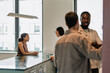 © Drobot Dean - A female employee sits at a table and holds a cup while talking to a female employee standing across from her while two male employees stand and talk next to them