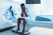 © Wavebreak Media - African man wearing hospital gown sitting on scanner table in scan room pressing hands to cheeks