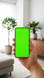 © MD Rashedul Islam - Hand holding mobile smartphone with blank green screen chroma key for mockup in bright living room interior with wooden cabinet plants and sofa background for home design app