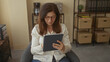 © Krakenimages.com - Woman in office holding tablet with hand on chin while seated amid shelves and storage boxes; concentration.
