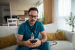 © Migma_Agency - Male doctor in scrubs using smart phone on living room sofa