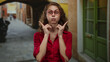 © Krakenimages.com - Young woman with glasses making funny expressions in a vibrant urban street with colorful buildings in the background, wearing a red shirt, conveying a playful mood.
