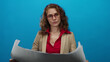 © Krakenimages.com - Woman examining large paper over isolated blue background wearing glasses and business attire, focused on content with professional demeanor.